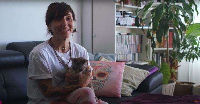 Mujer sosteniendo un gato en un sofá con una almohada de calavera y plantas en el fondo.