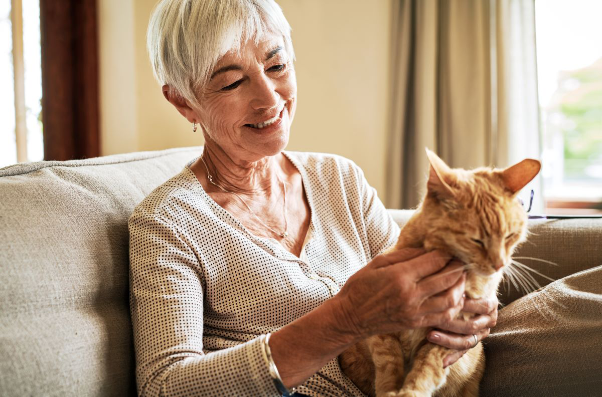Femme âgée souriante caressant un chat sur un canapé.