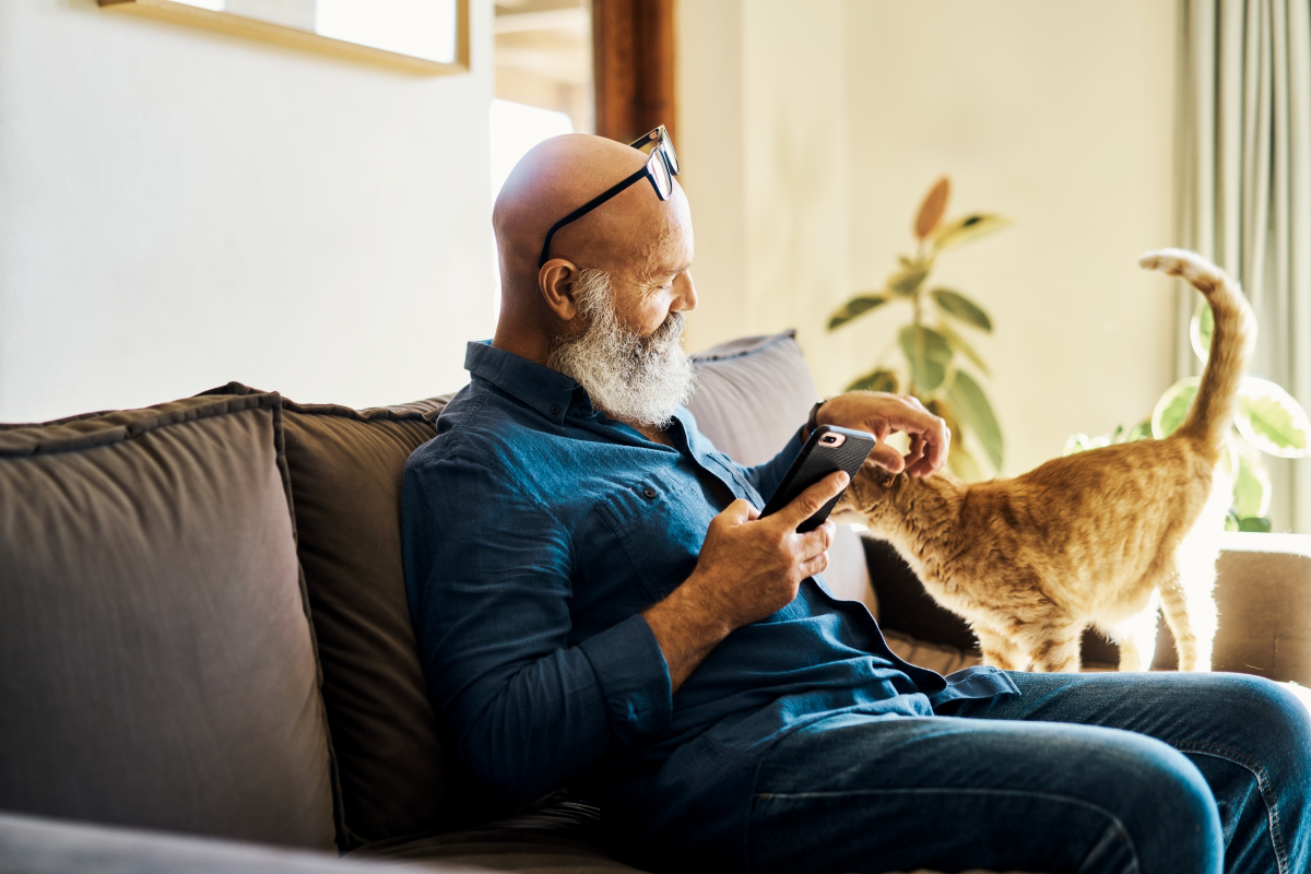 Man petting a cat on a sofa while holding a smartphone.