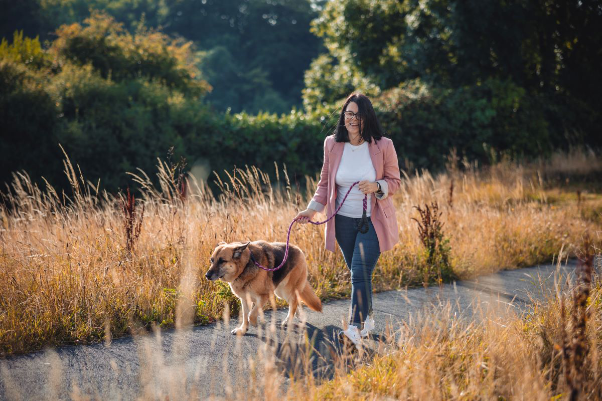Woman walking a dog on a path through a field on a sunny day.